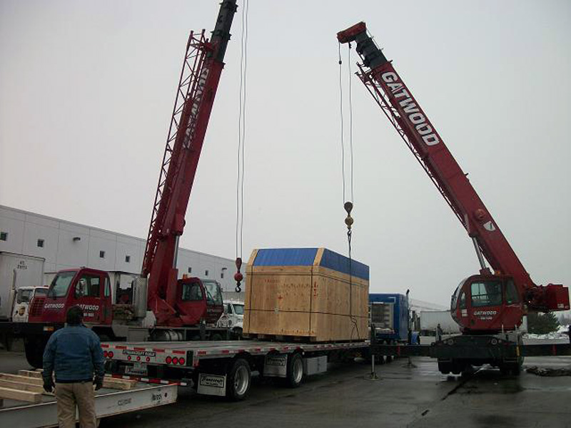 Large wooden box lifted by two cranes and placed on large semi-truck bed for delivery